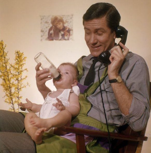 Telefonerende vader met huishoudschort geeft baby de fles. [1961].